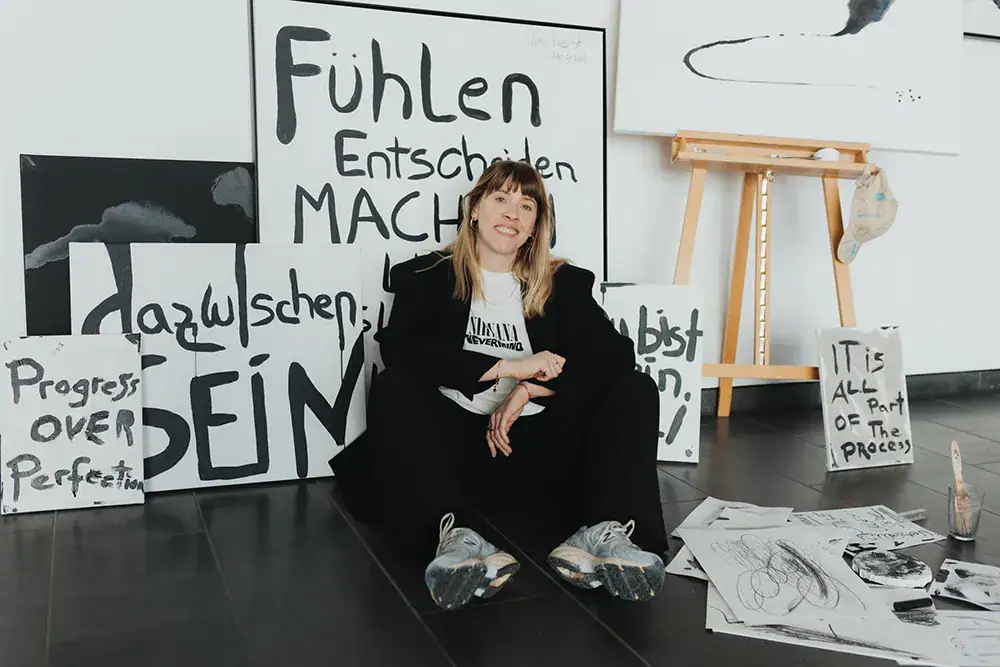 Künstlerin sitzt vor inspirierenden Schwarz-Weiß-Textkunstwerken, umgeben von Malutensilien, in eine.