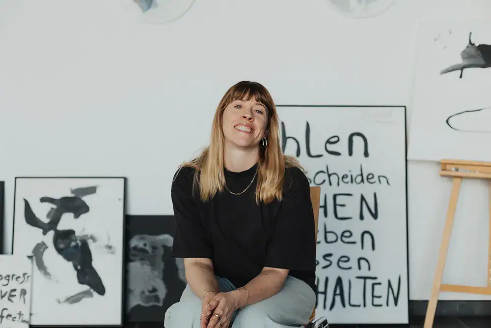 Frau sitzt vor Kunstwerken in einer Galerie, lächelt freundlich, kreative Atmosphäre, moderne Kunst,.